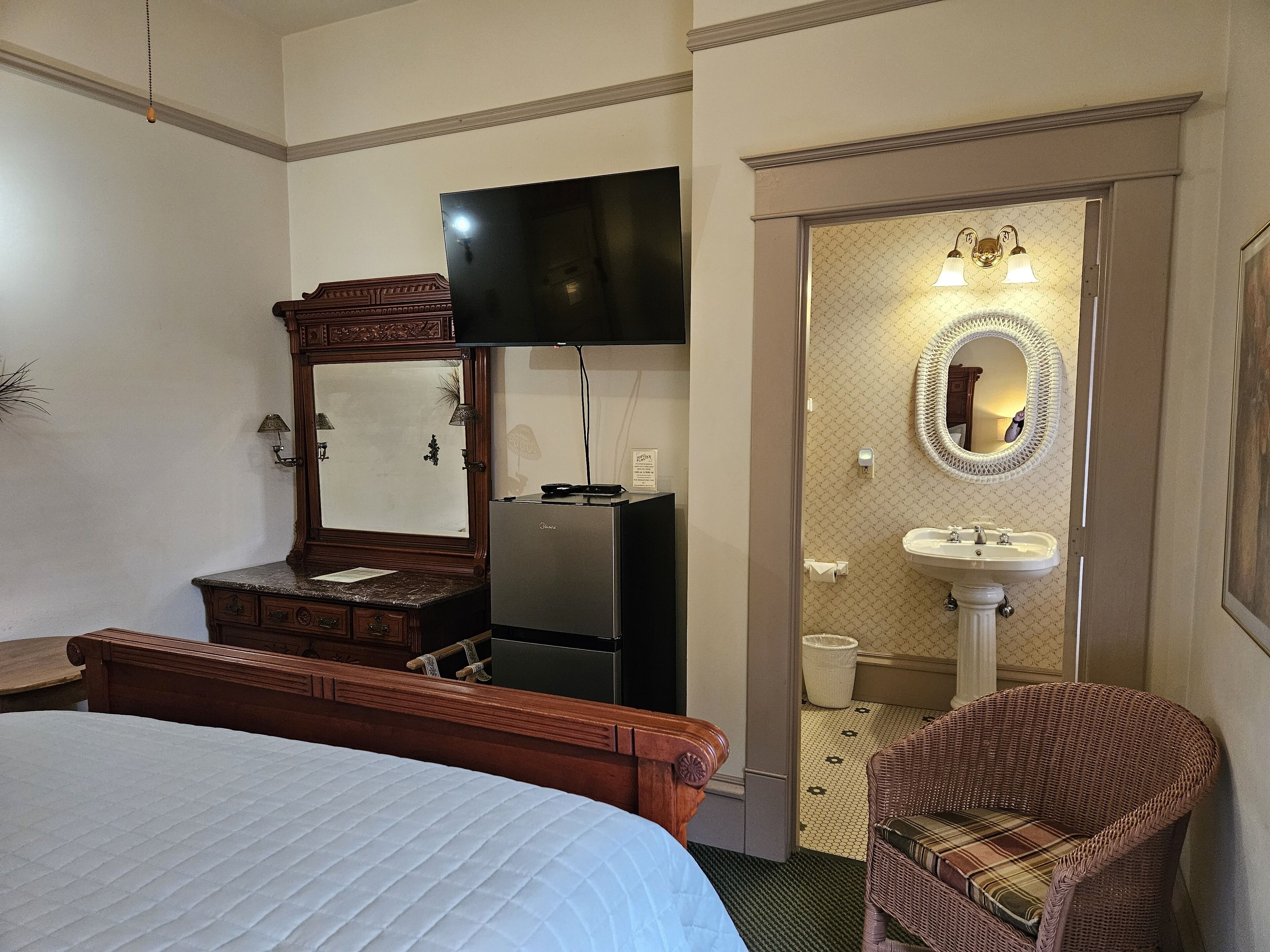 A hotel room with a bed, a wooden dresser, a flat-screen TV, a refrigerator, a wicker chair, and a bathroom with a sink and mirror.