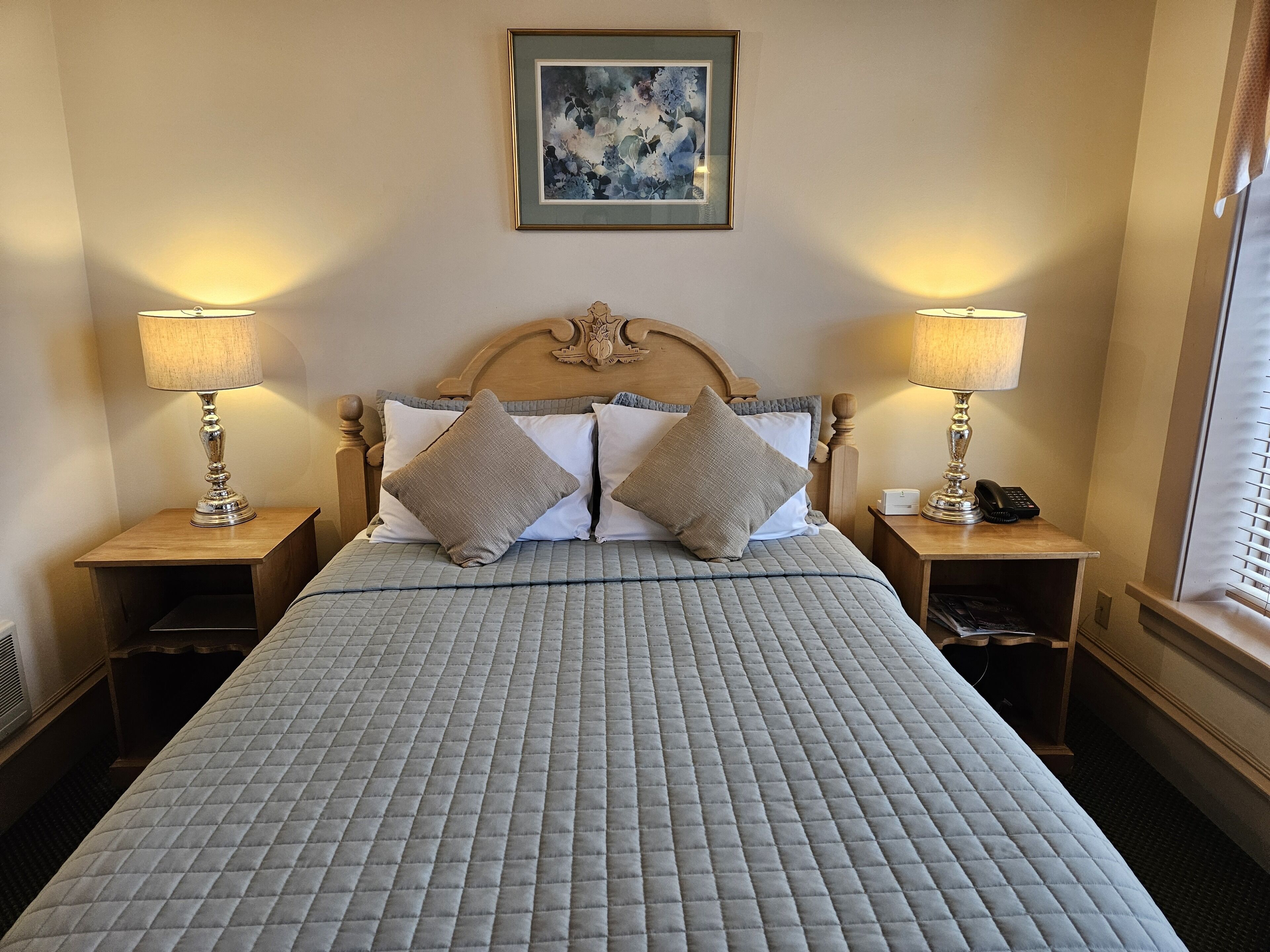 A neatly made bed with a quilted grey bedspread, two bedside lamps, and a framed floral artwork above the headboard.