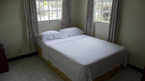 A single bed with white bedding in a room with tiled flooring and two windows with curtains. - Malfranza Apartments (Bridgetown)