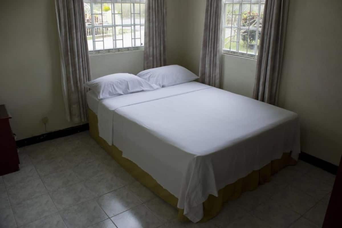 A single bed with white bedding in a room with tiled flooring and two windows with curtains.