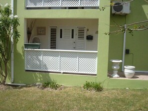 Edificio de dos pisos con fachada verde, un balcón con barandilla de celosía blanca y un pequeño jardín con una urna blanca y una maceta.