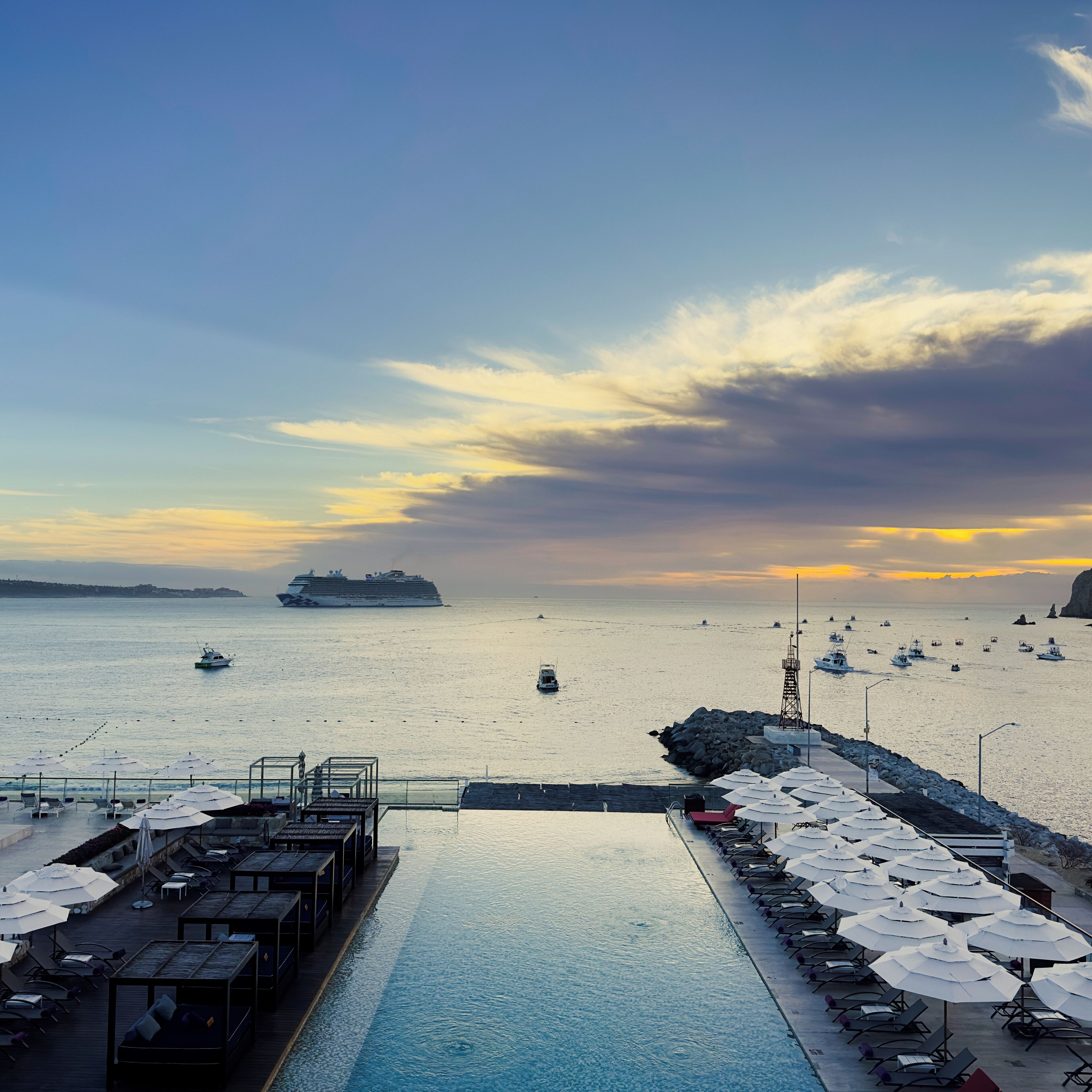 A waterfront pool area with lounge chairs and umbrellas, a pier with boats, and a cruise ship in the distance.
