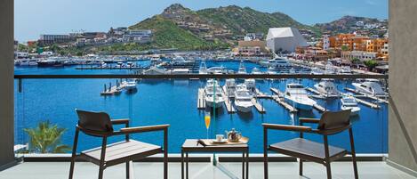 Un balcón con dos sillas y una mesa, con vistas a un puerto deportivo con barcos y montañas al fondo.