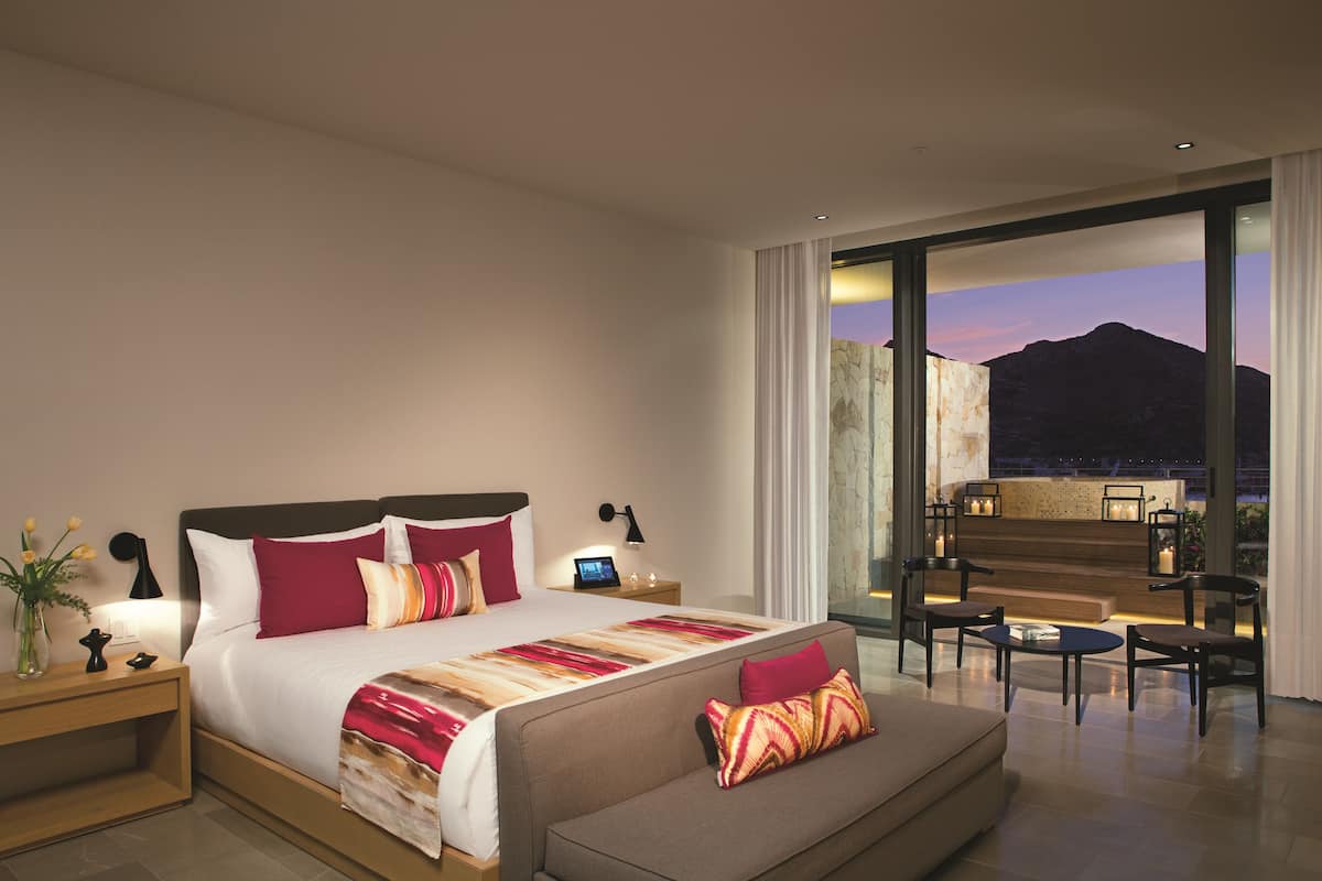 Habitación de hotel moderna con una cama grande, un sofá, un escritorio y vistas a montañas al anochecer.