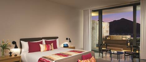 A modern hotel room with a large bed, a sofa, a desk, and a view of mountains at dusk.