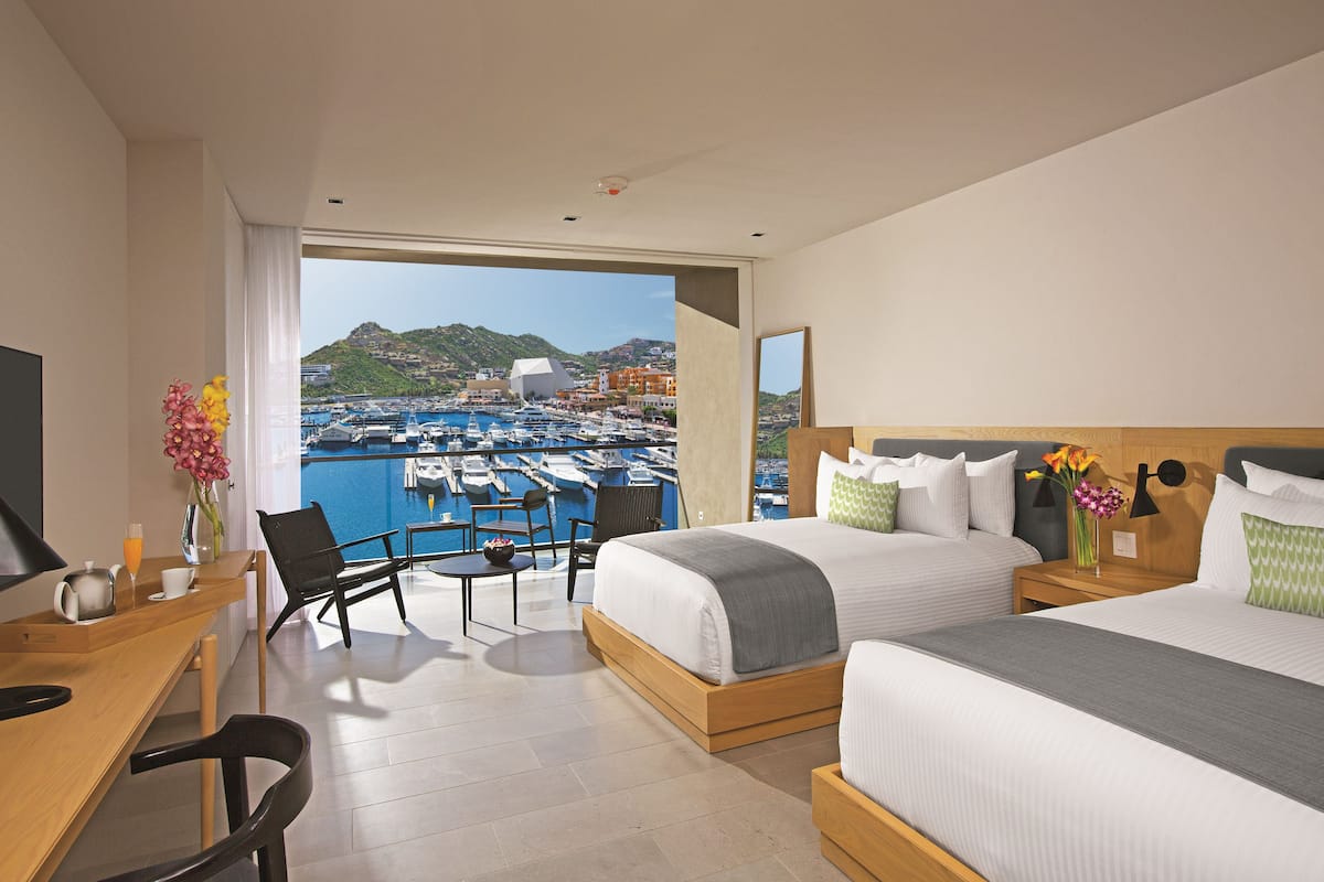 Habitación de hotel con dos camas, un escritorio y una silla. Se ve un puerto deportivo con barcos y edificios.