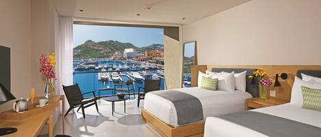Habitación de hotel con dos camas, un escritorio y una silla. Se ve un puerto deportivo con barcos y edificios.