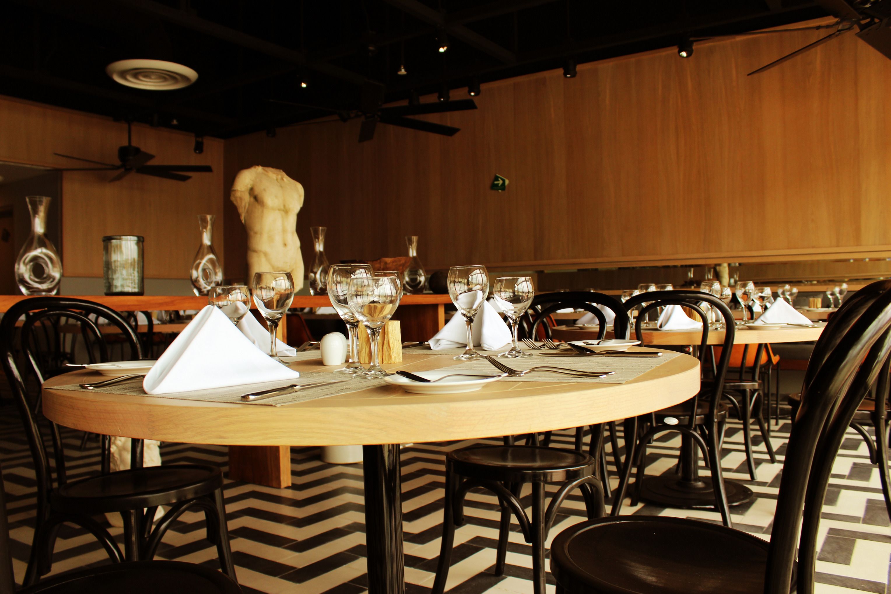 A restaurant with round tables set for dining, chairs, and a wooden wall with a ceiling fan.