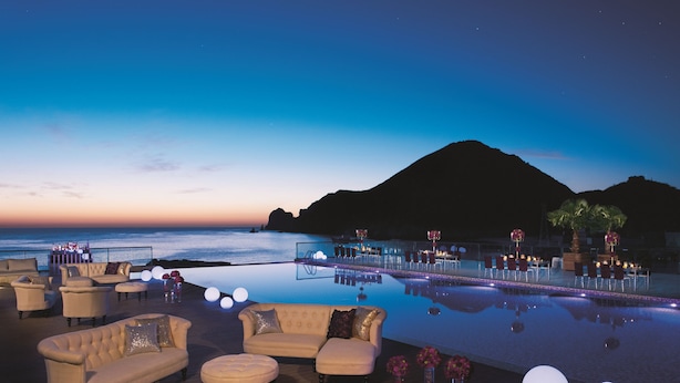 A poolside area with lounge chairs and a bar, overlooking a body of water and a mountain.