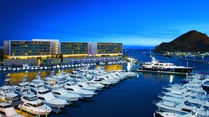 A marina with numerous yachts docked, a large hotel building with multiple floors, and a prominent mountain in the background.