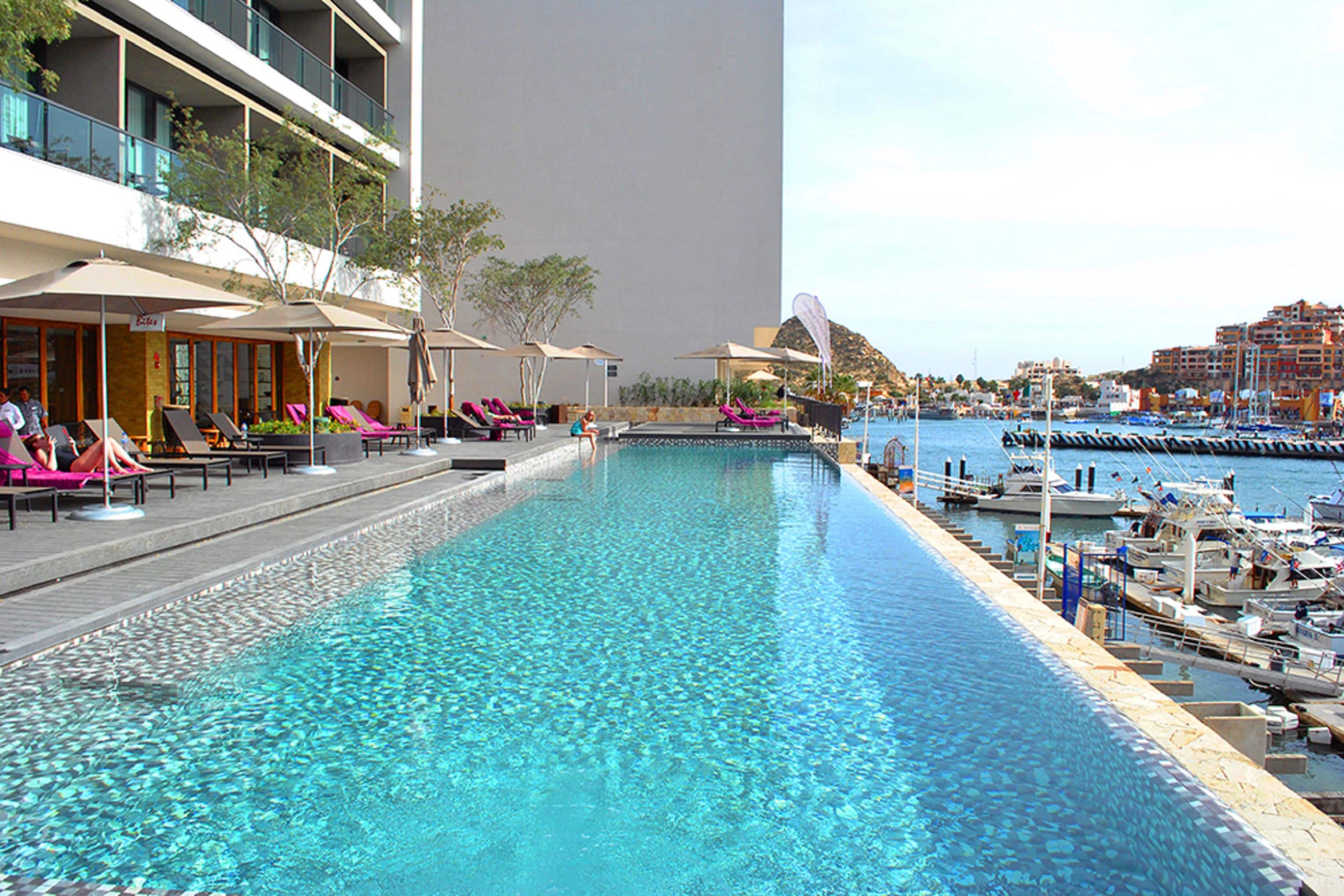 A swimming pool with lounge chairs and umbrellas, a marina with boats, and a building with balconies overlooking the pool.