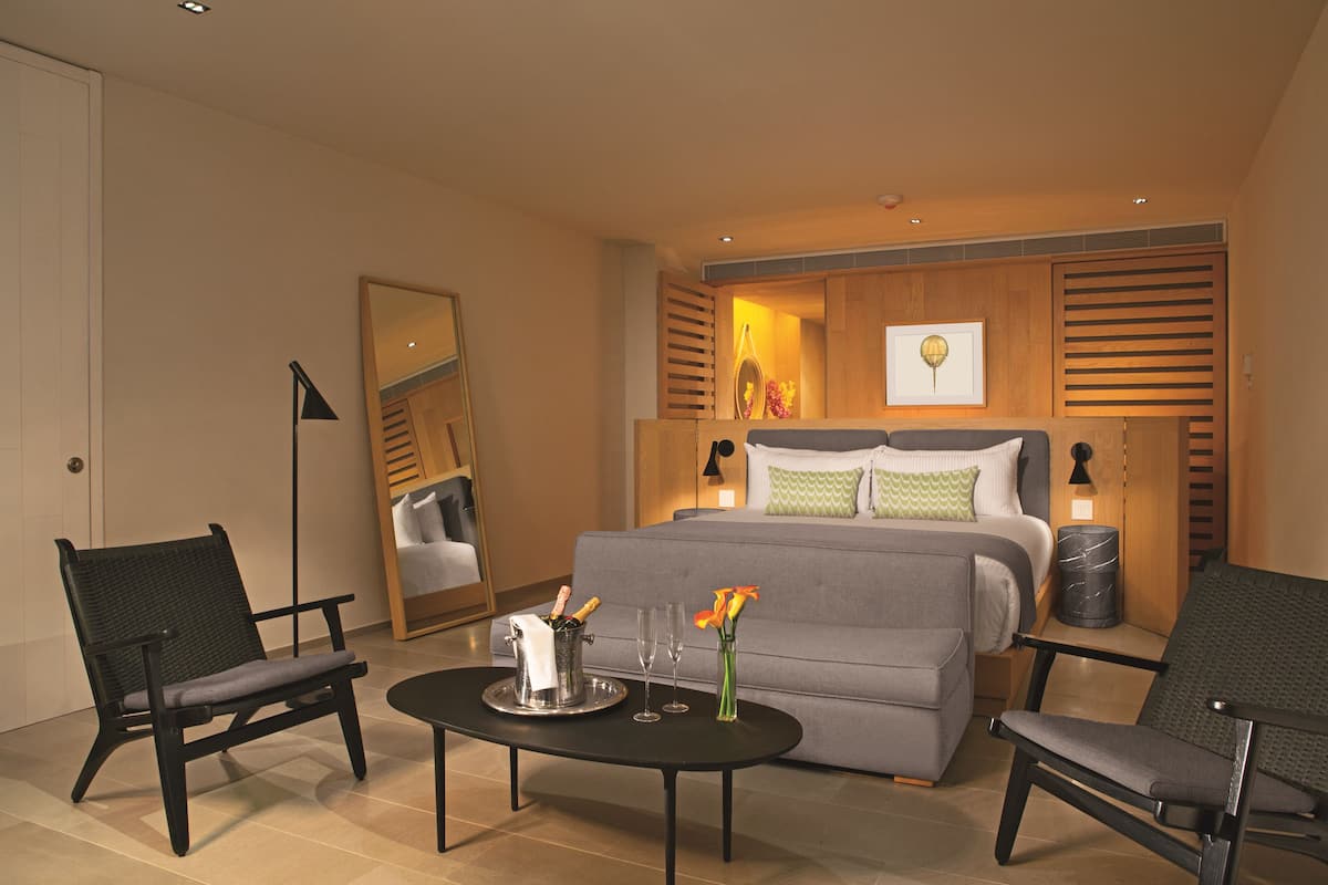 Una habitación de hotel moderna con cama, dos sillones, una mesita de centro y una mesita de noche con lámpara.