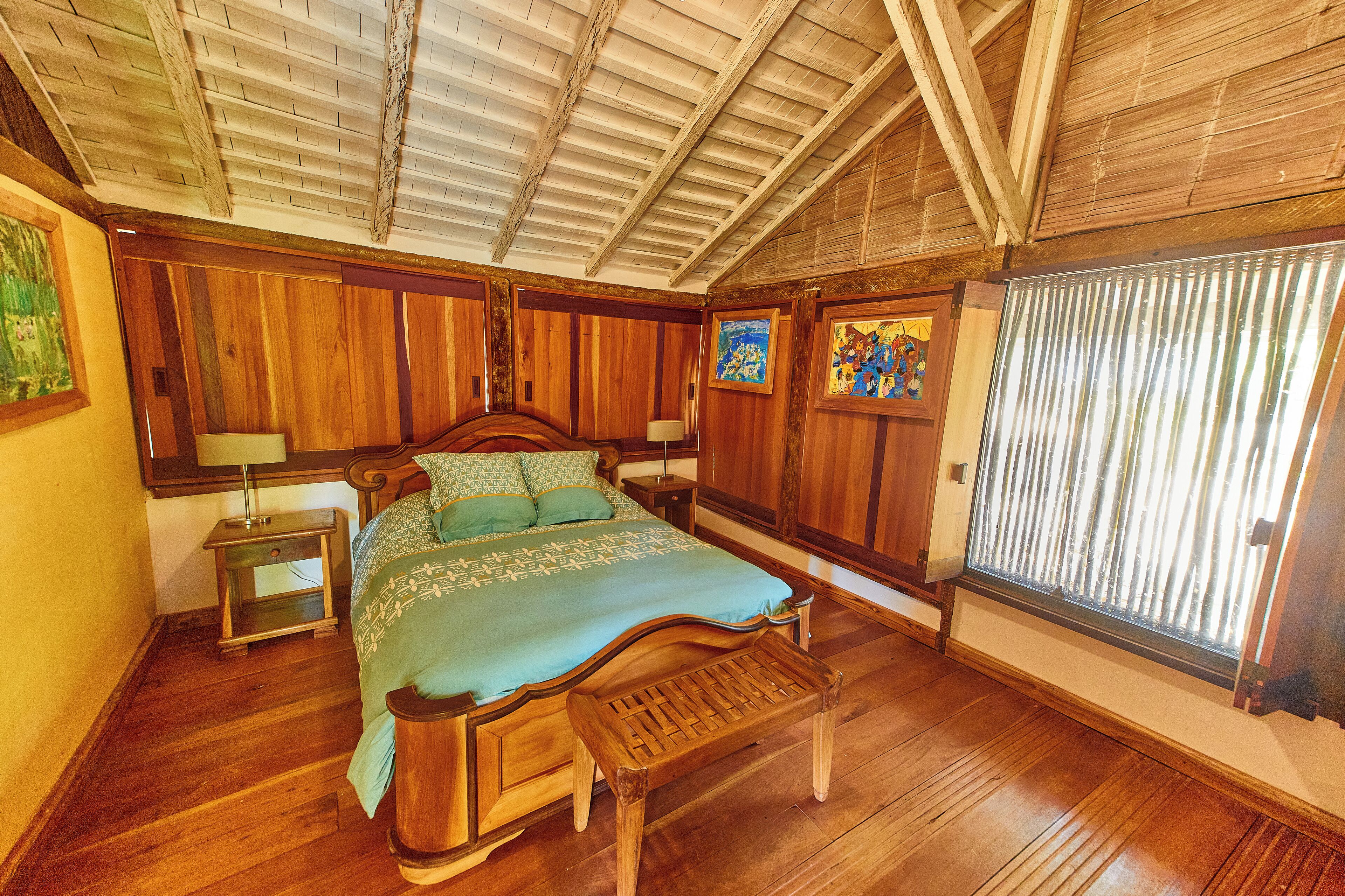 A wooden-framed bedroom with a bed, bench, nightstands, and artwork on the walls.