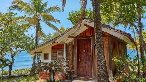 Una casa de madera con puerta roja, rodeada de palmeras y vista despejada al océano.