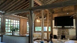 Un interior rústico con vigas de madera a la vista, una chimenea de piedra, un televisor y una zona de estar con cojines estampados.