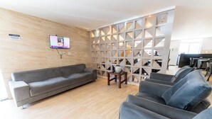 Een moderne woonkamer met een grijze bank, een aan de muur bevestigde televisie en een decoratieve scheidingswand met geometrische patronen.