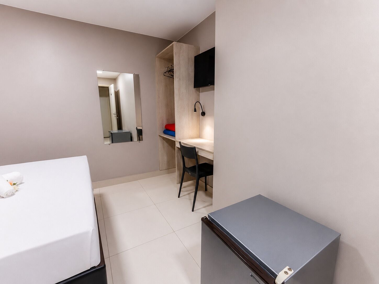 A compact hotel room with a bed, a desk, a chair, a TV, and a small refrigerator.