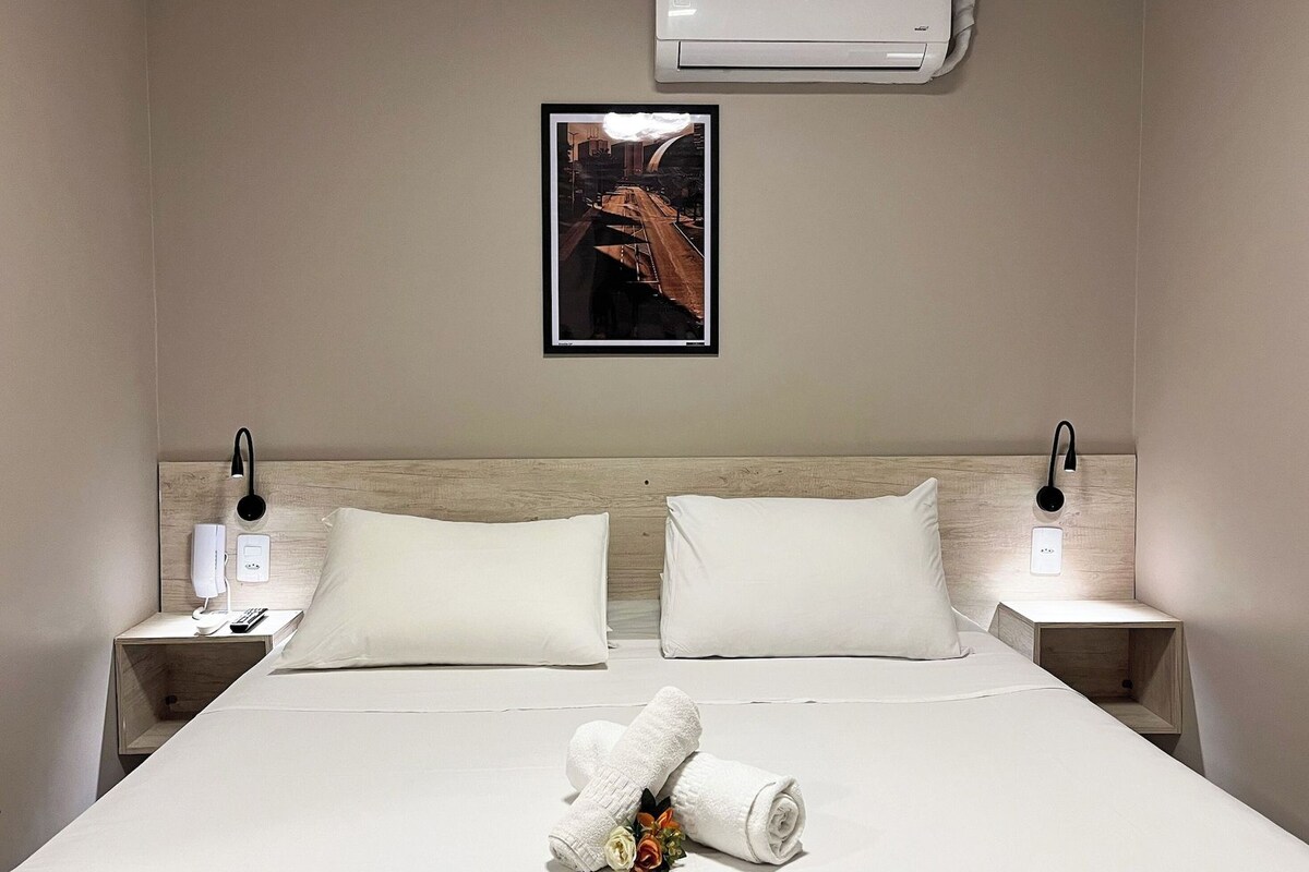 A hotel room with a bed, two bedside tables, an air conditioner, a framed picture on the wall, and a rolled towel with flowers on the bed.