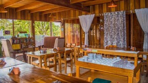 A rustic wooden interior with multiple wooden tables and chairs, a bookshelf, and a view of greenery outside. - Hotel Holístico Monteverde (Monteverde)