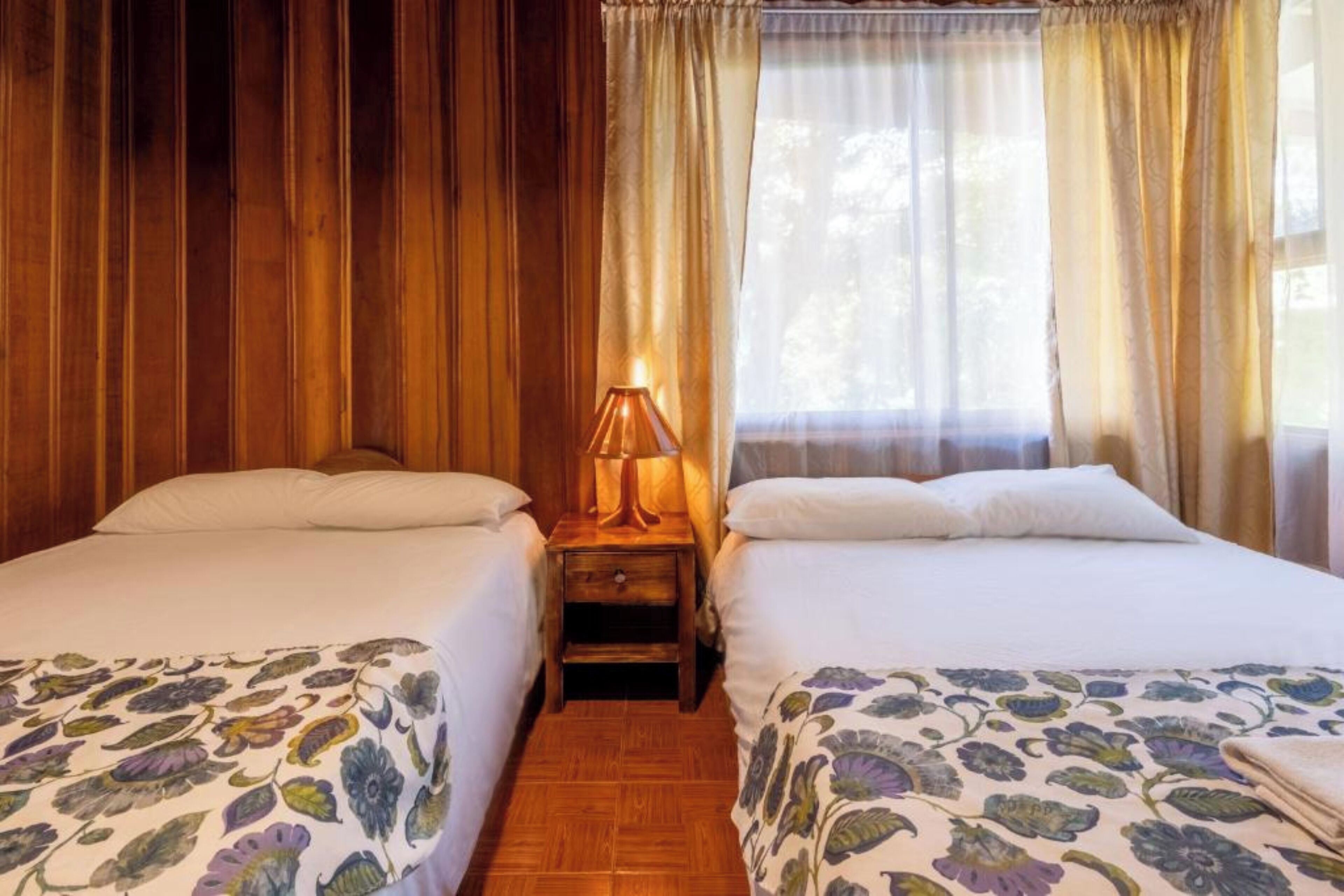 A room with two beds, wooden paneling on the walls, a nightstand with a lamp, and sheer curtains.