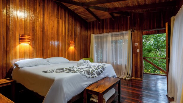 A wooden room with a bed, two wall-mounted lights, a bench, and a door leading to an outdoor area with greenery. - Hotel Holístico Monteverde (Monteverde)