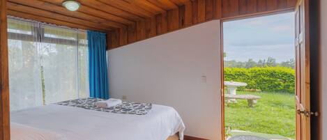 Une chambre avec un plafond en bois, un lit recouvert de draps blancs, une fenêtre avec des rideaux bleus et une vue sur un jardin.