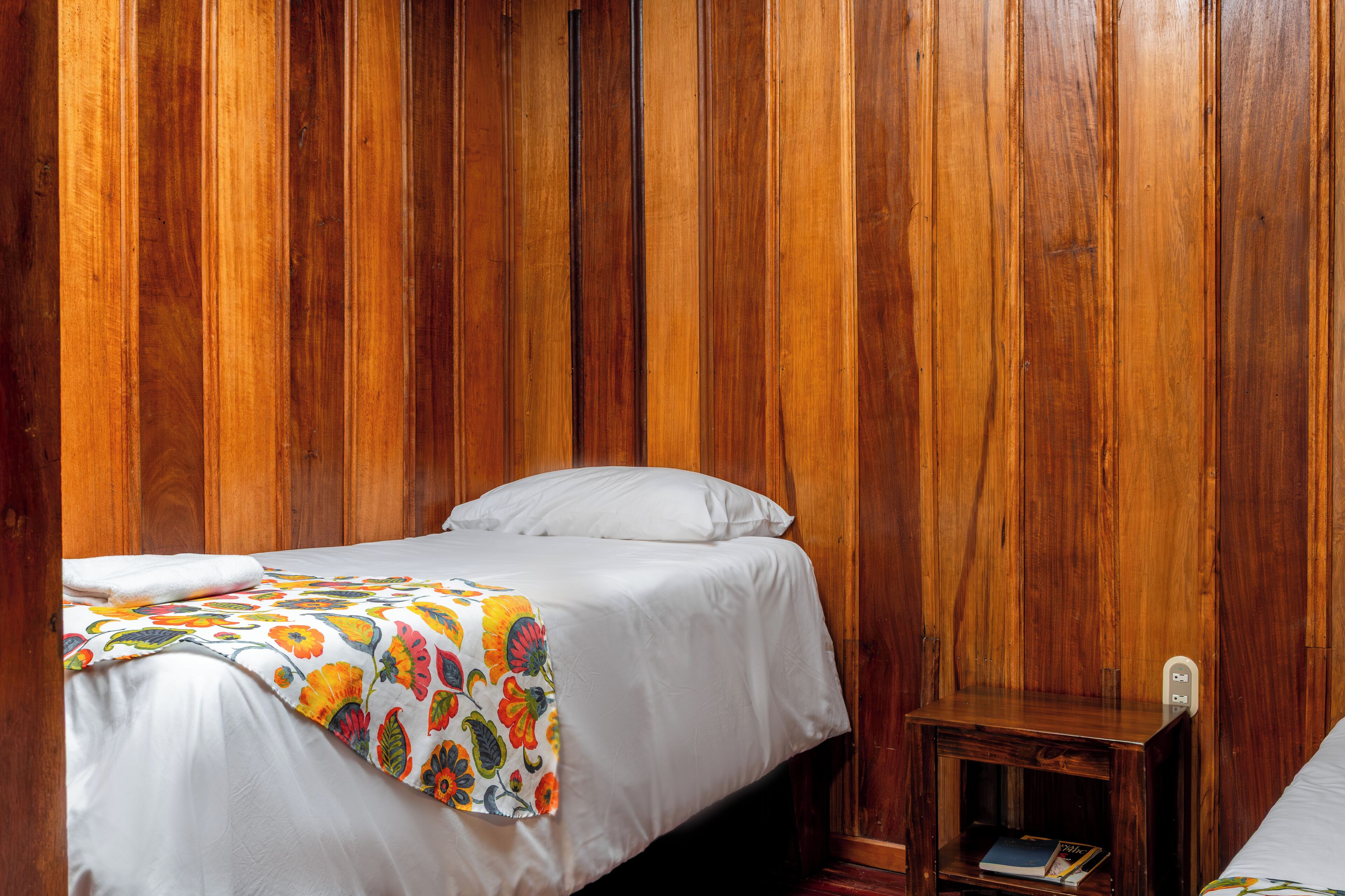 Un lit simple avec un drap blanc et une couverture à motifs floraux colorés, une table de chevet en bois avec un livre, et un lambris en bois.