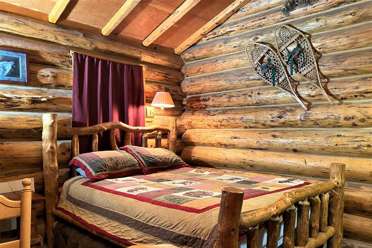 A log cabin bedroom with a wooden bed, a quilt, a wooden headboard, a table, a chair, a lamp, and a framed picture on the wall.