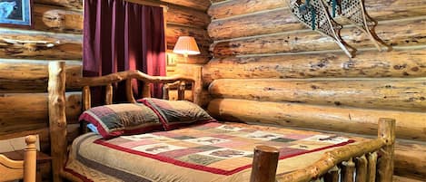 Un dormitorio de cabaña de madera con una cama de madera, colcha, cabecera de madera, una mesa, una silla, una lámpara y un cuadro en la pared.