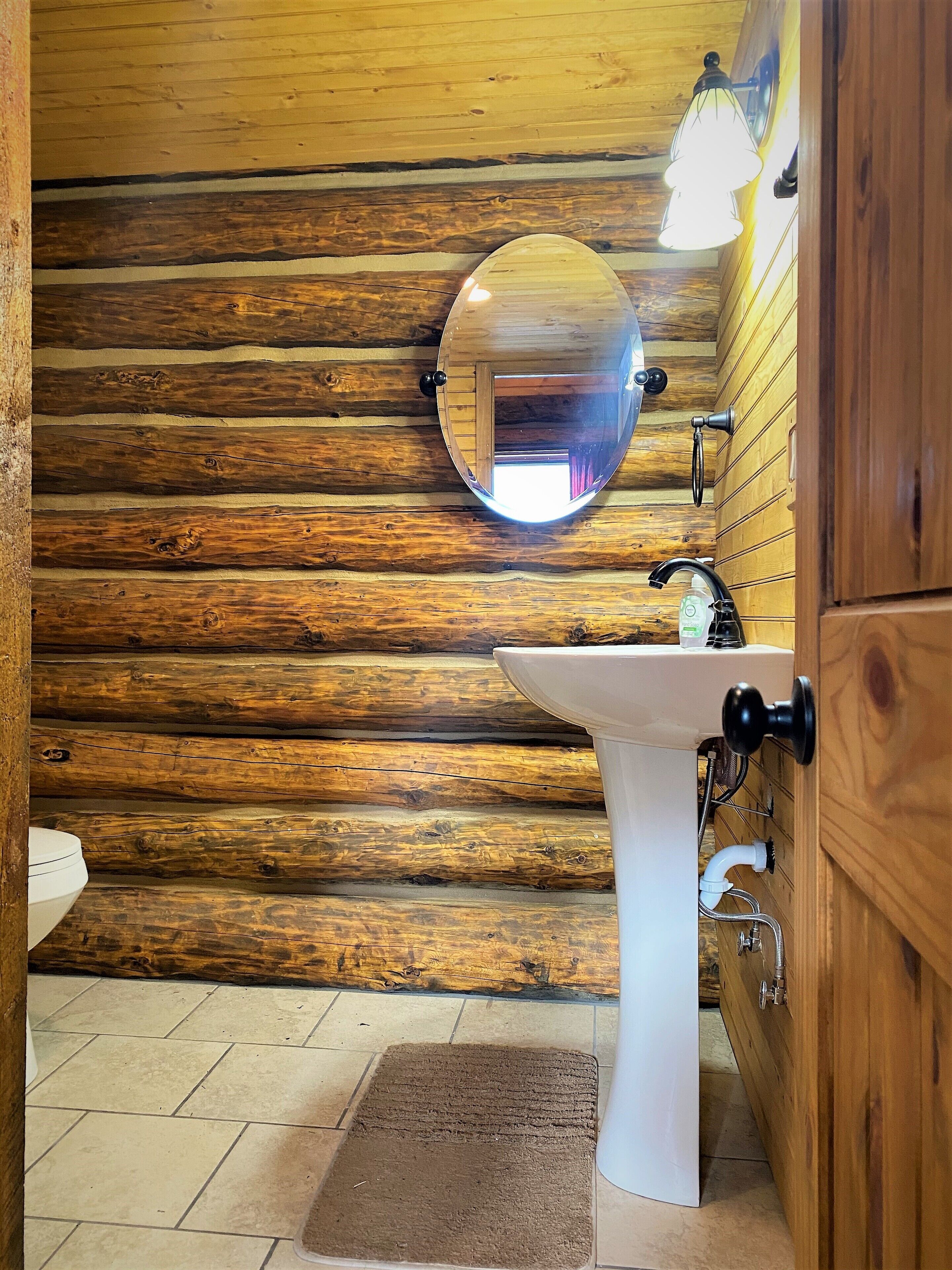 Un baño rústico con pared de madera, un espejo redondo, un lavamanos blanco y una alfombra marrón.