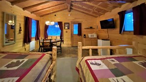 Acogedora habitación tipo cabaña de madera con techo de madera, una mesa de comedor con sillas, dos camas con colchas, un televisor y una ventana con cortinas.