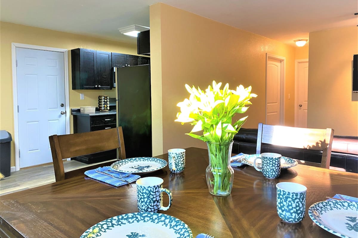 A dining area with a wooden table set for four, surrounded by chairs. A vase with flowers is placed in the center.