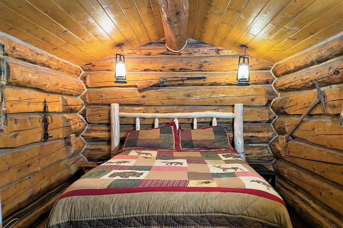 A log cabin bedroom with a bed, two wall-mounted lanterns, and a wooden ceiling.