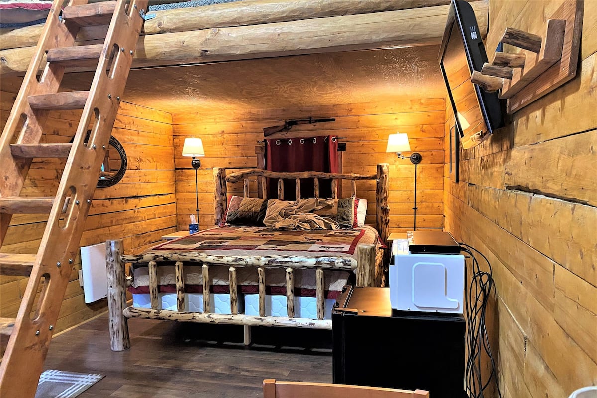 A wooden cabin bedroom with a bed, two bedside lamps, a wooden ladder, a desk, and a chair.