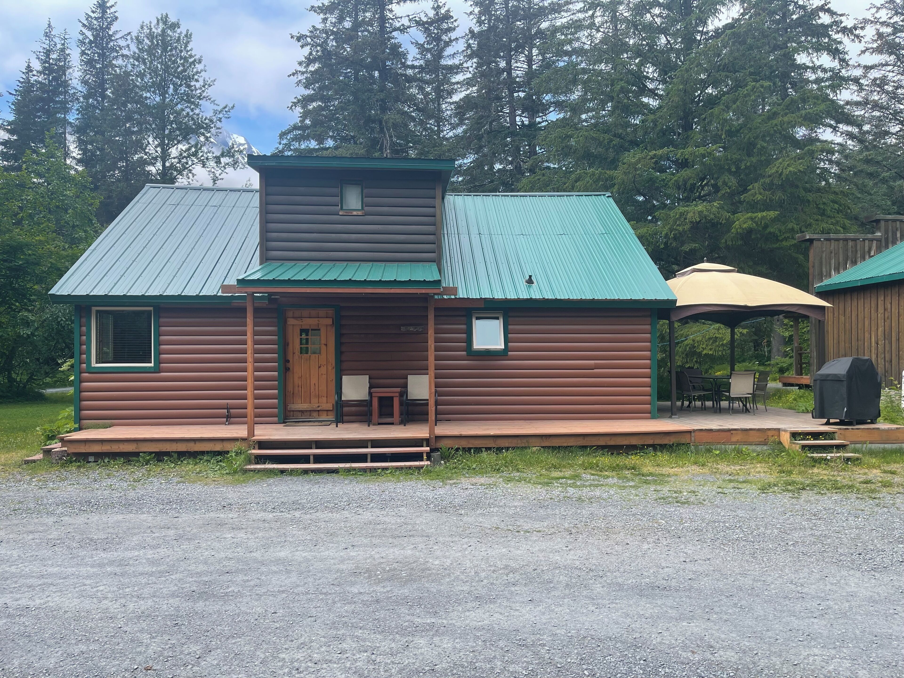 A rustic cabin with a green metal roof, a front porch with two chairs, and a covered patio area.