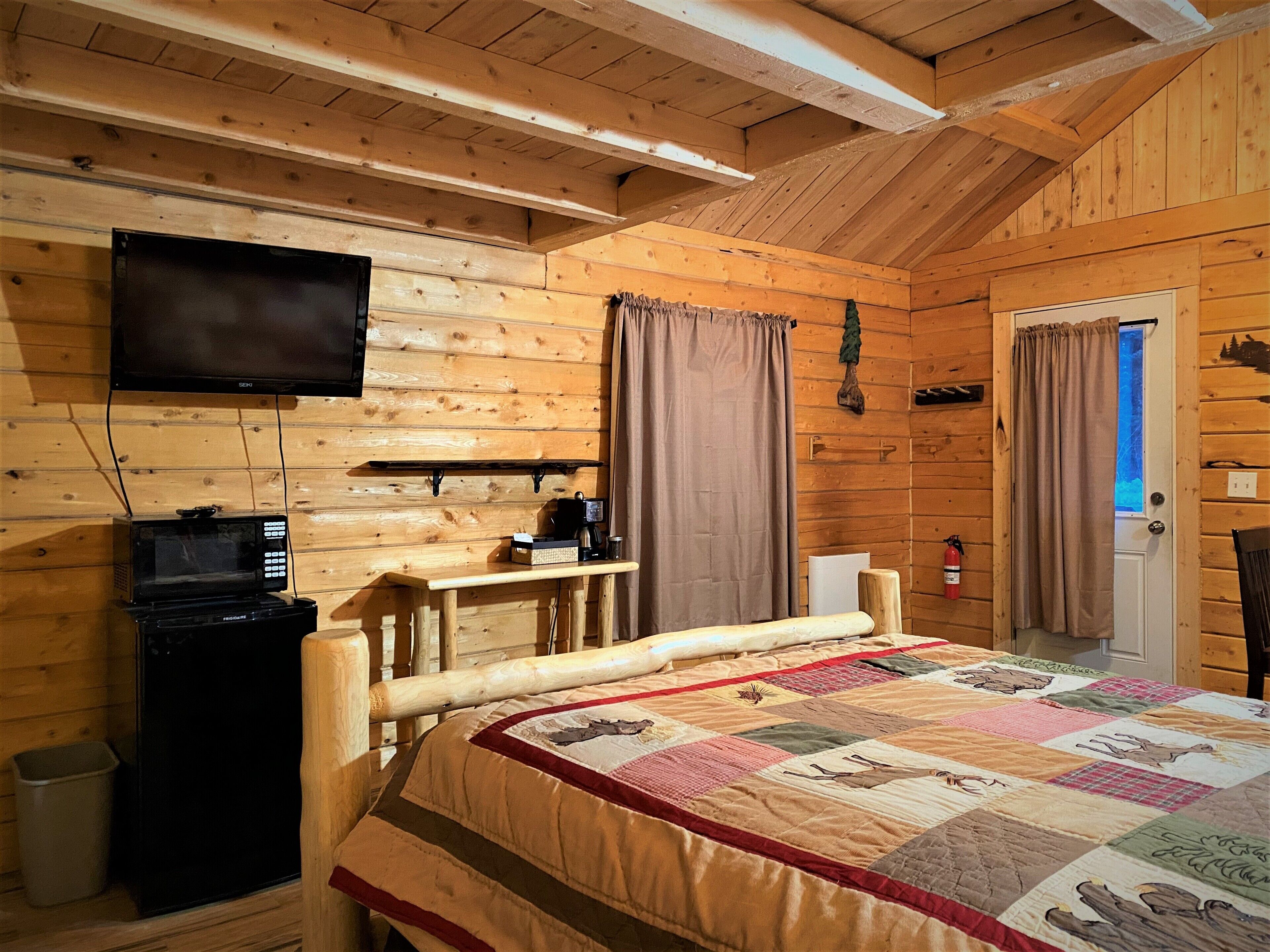 Une chambre confortable de type chalet, avec un lit, un téléviseur fixé au mur, une petite kitchenette équipée d’un four à micro-ondes et une fenêtre avec des rideaux.