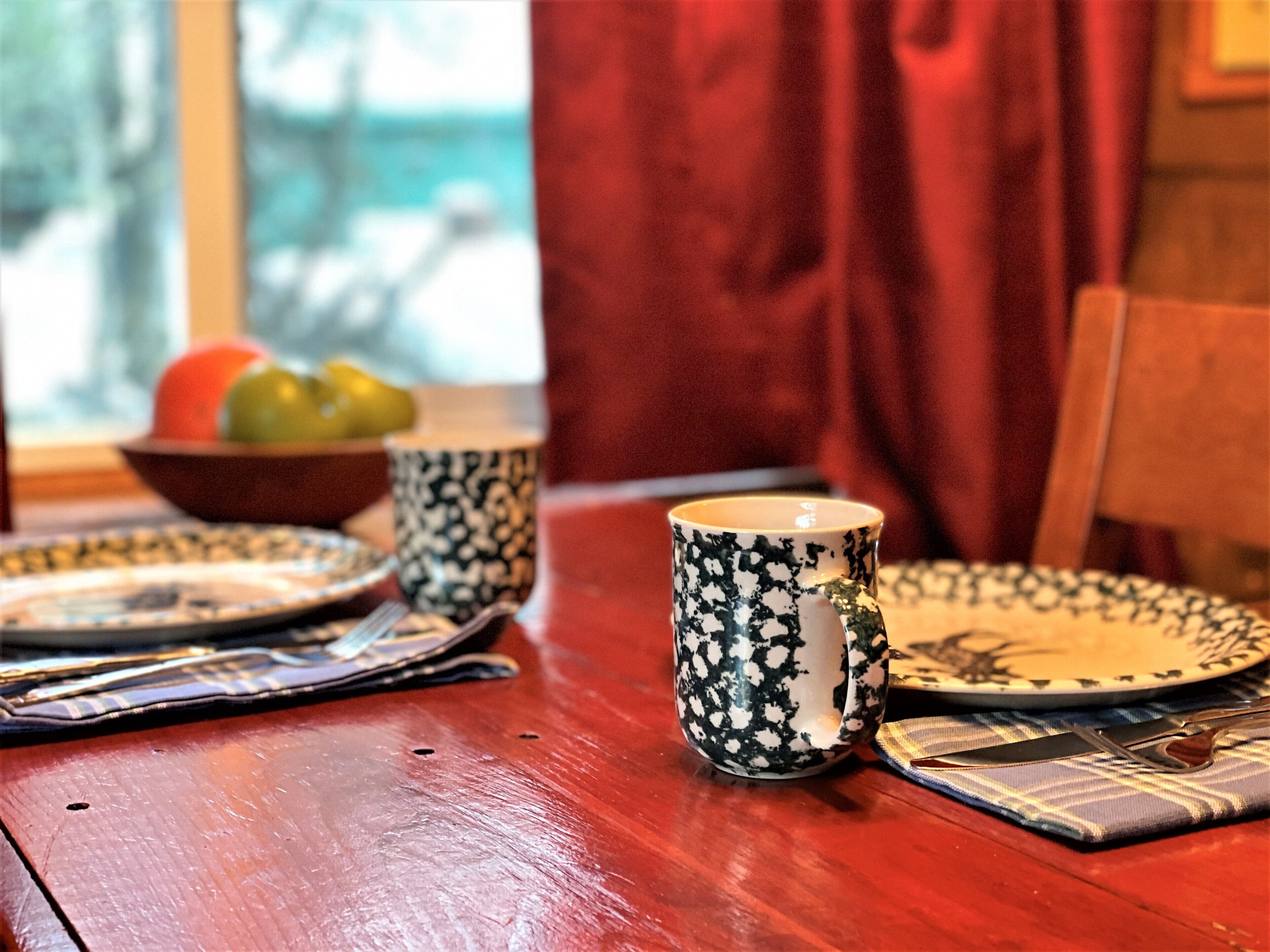 A wooden dining table with a bowl of fruit, two patterned mugs, a plate, and cutlery.