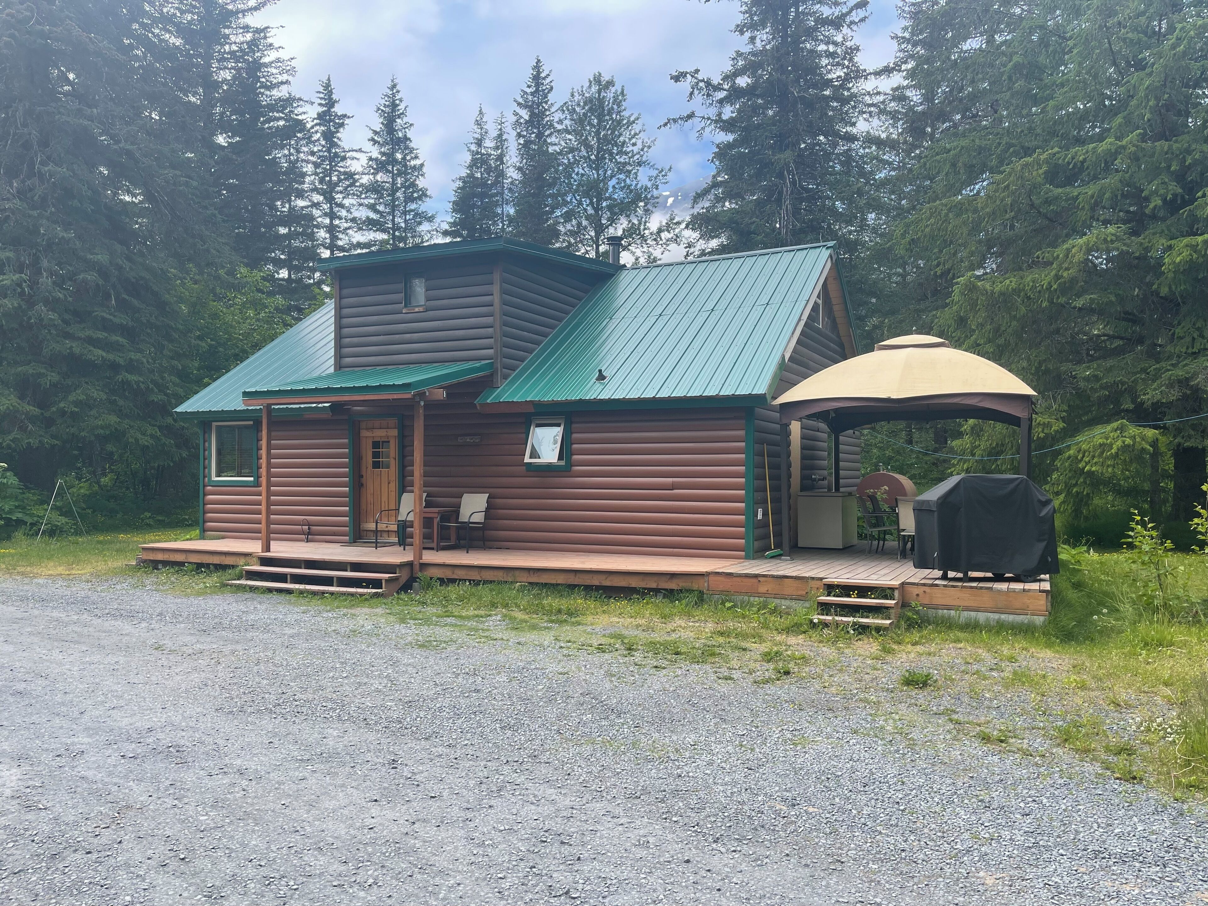 A rustic cabin with a green roof, a porch with seating, and a barbecue area.