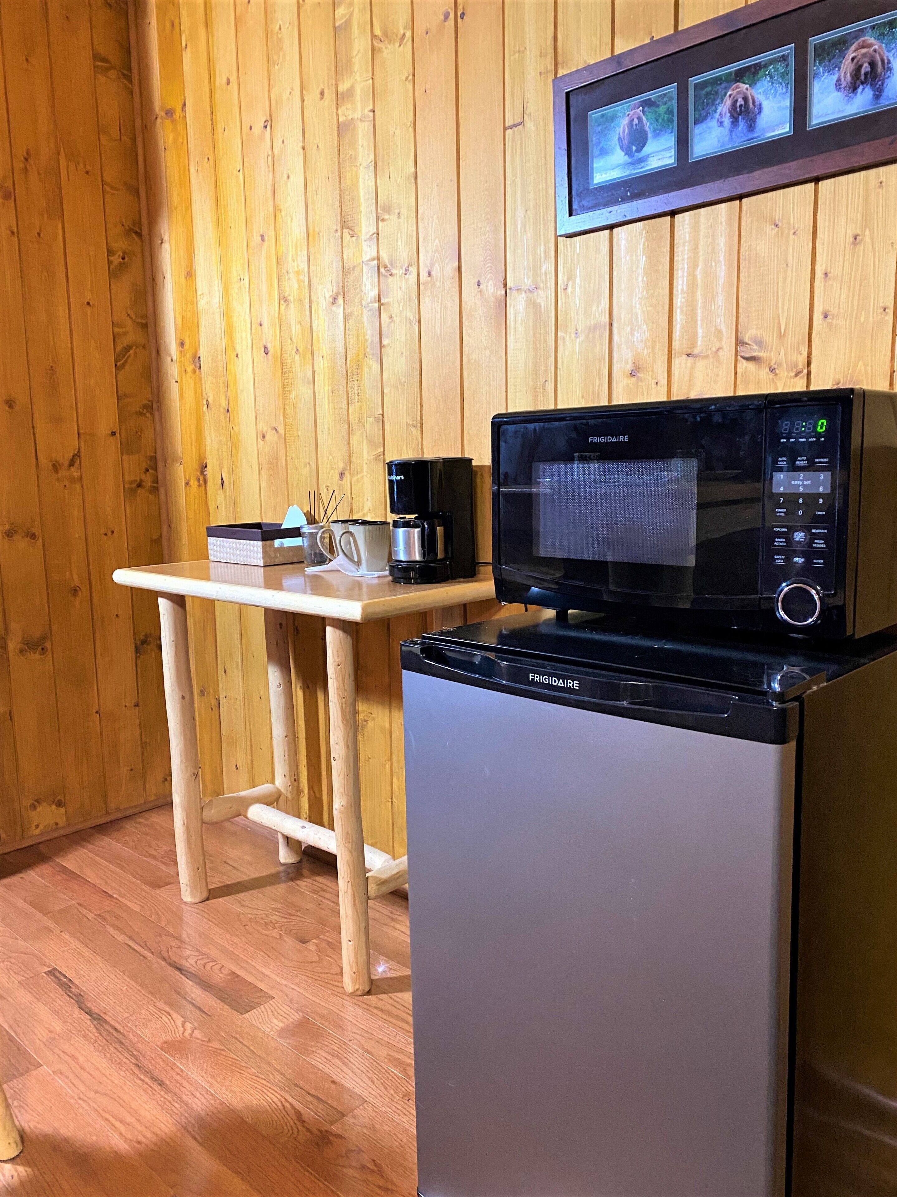 A compact kitchen area with a Frigidaire microwave and refrigerator, a wooden table with a coffee maker, and a wall-mounted picture frame displaying three bear images.