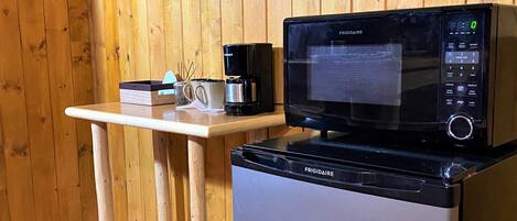 A compact kitchen area with a Frigidaire microwave and refrigerator, a wooden table with a coffee maker, and a wall-mounted picture frame displaying three bear images.