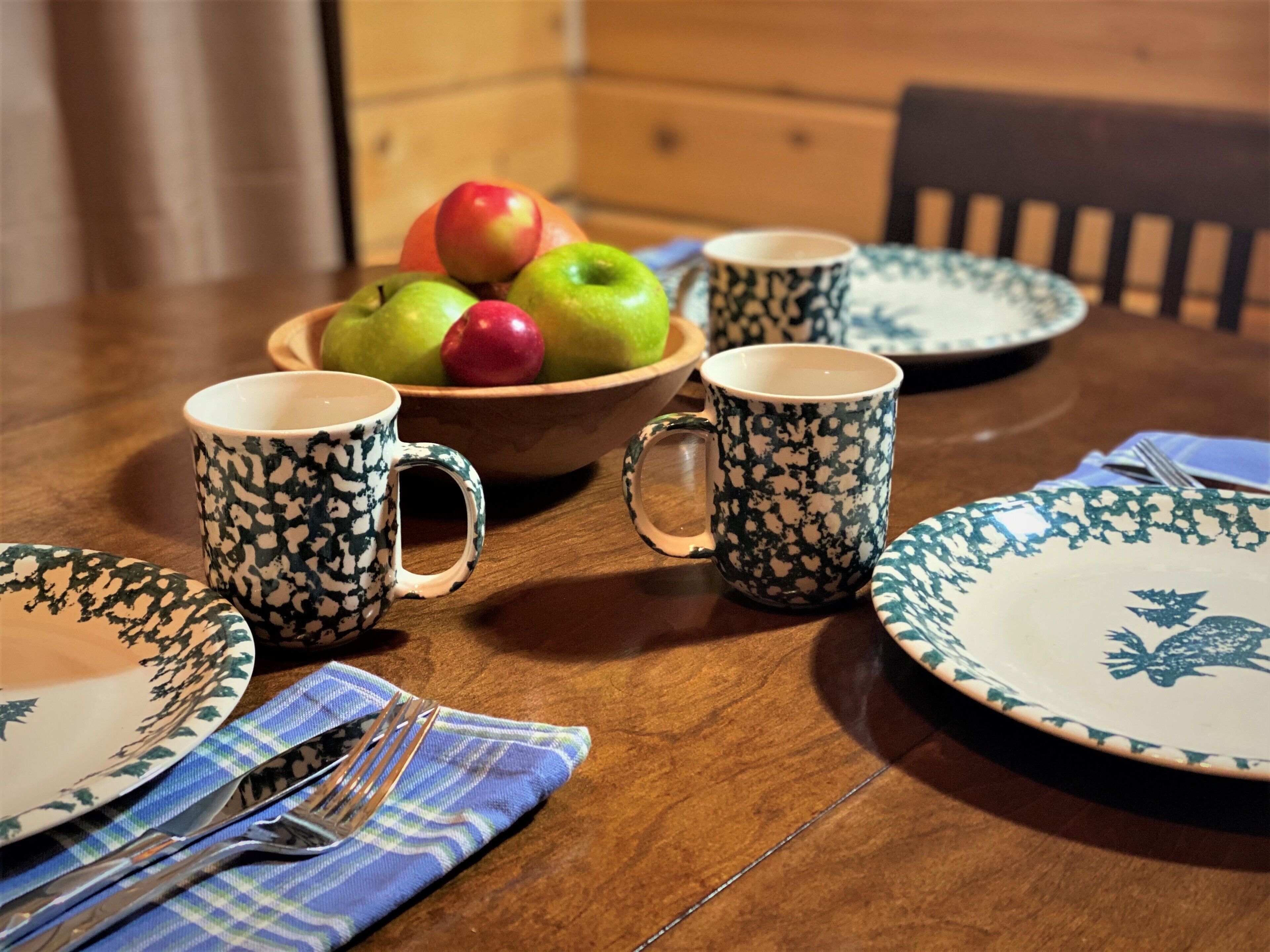 A wooden dining table with a bowl of fruit, two patterned mugs, and a blue napkin with a fork.