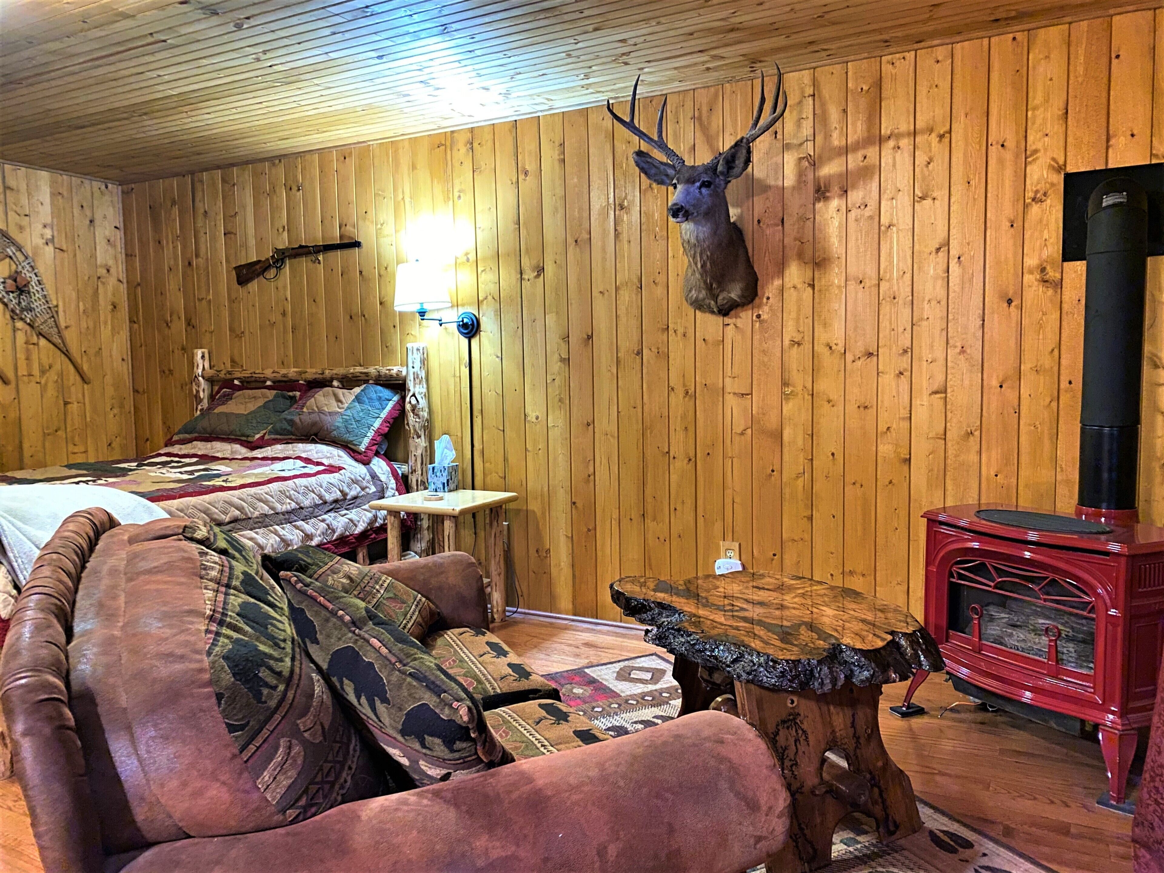 A cozy room with a bed, a leather couch, a wooden table, a mounted deer head, and a red wood-burning stove.