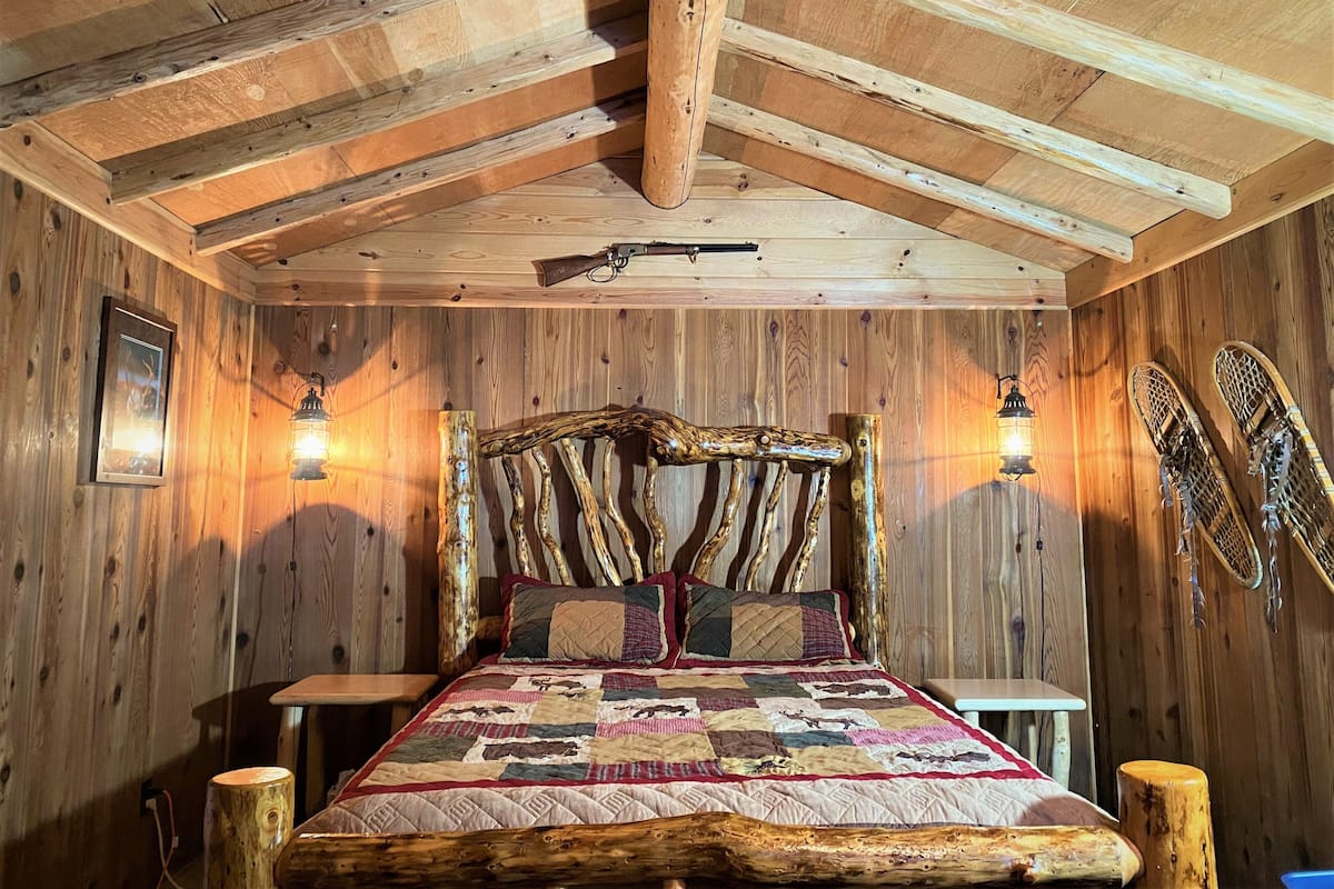 A rustic wooden bedroom with a bed, two wall-mounted lights, and a rifle on the ceiling.