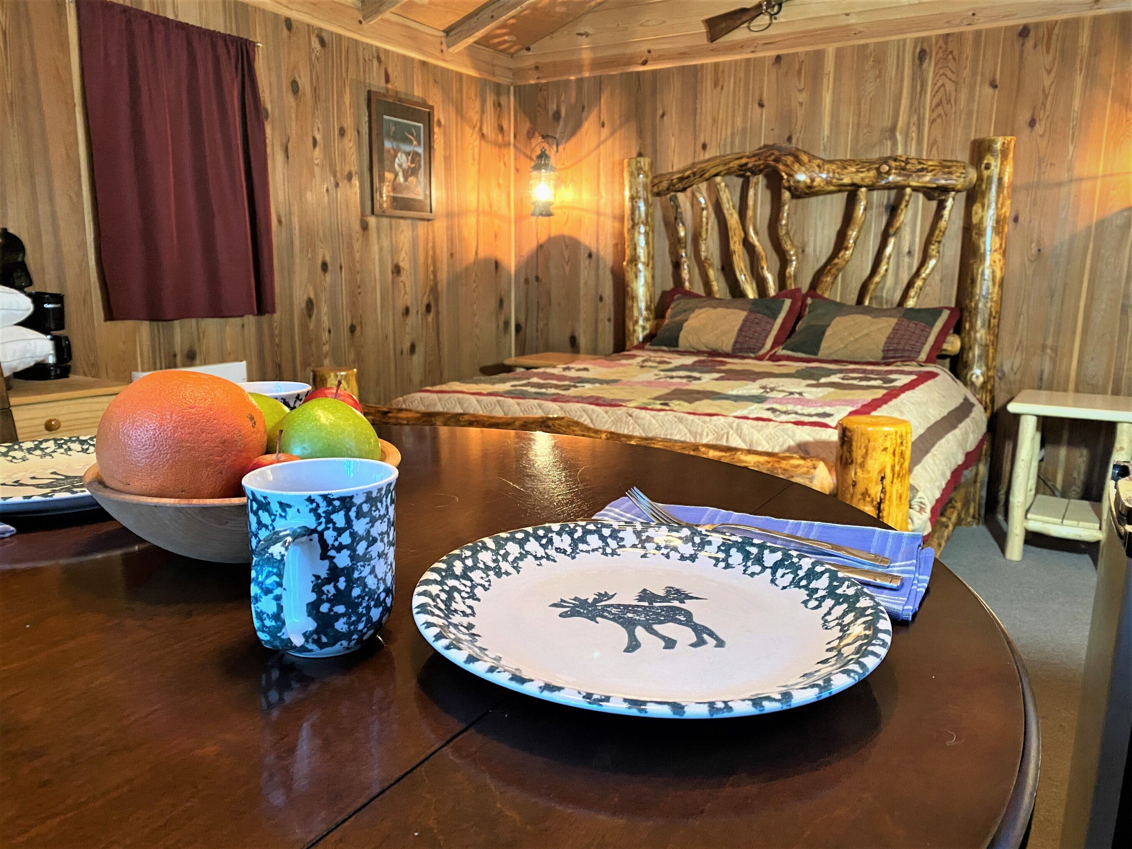 A wooden room with a bed, a table with fruit and plates, and a wall-mounted picture.