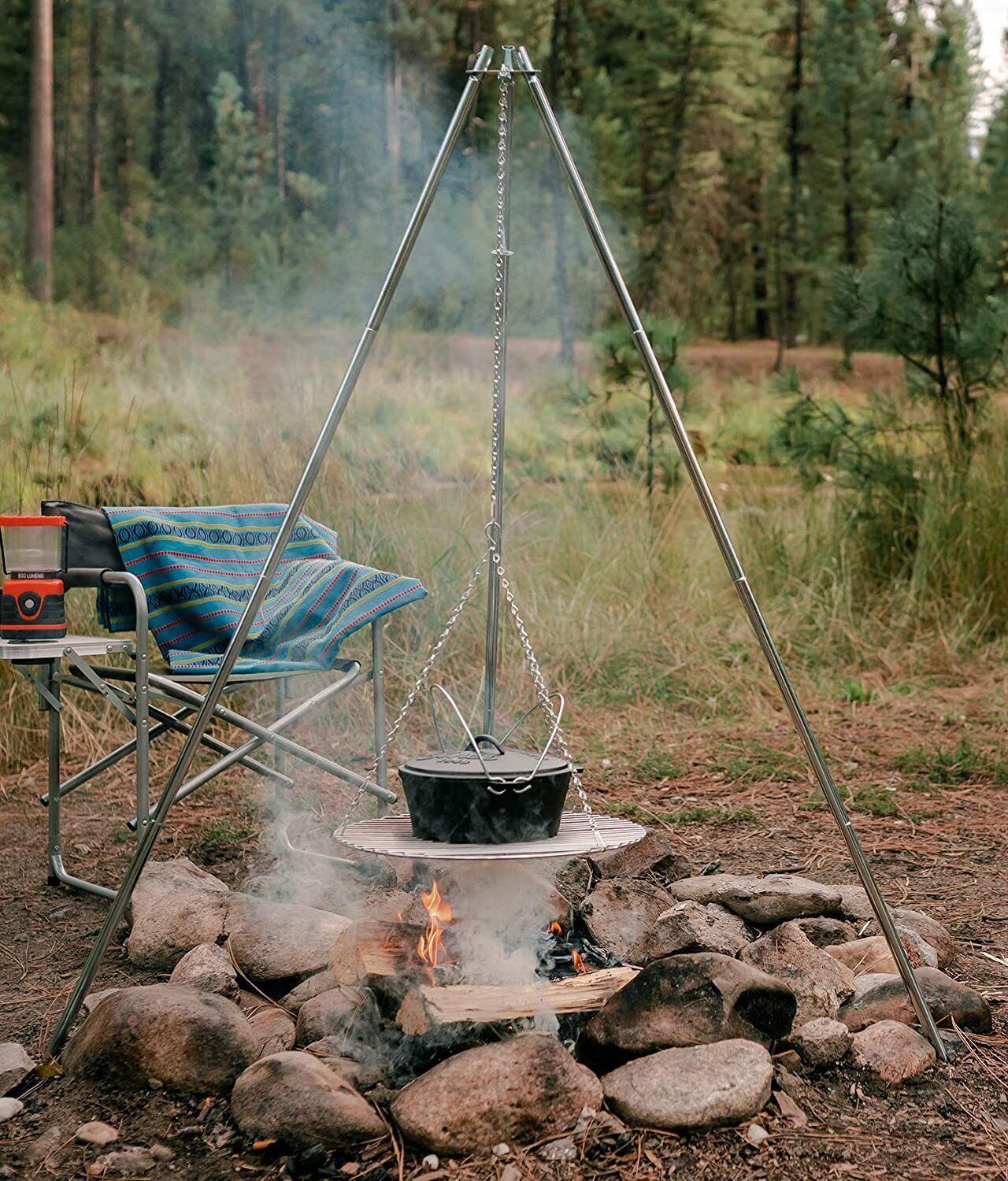 A portable camping stove with a pot on a metal stand, surrounded by rocks and a chair with a blanket.