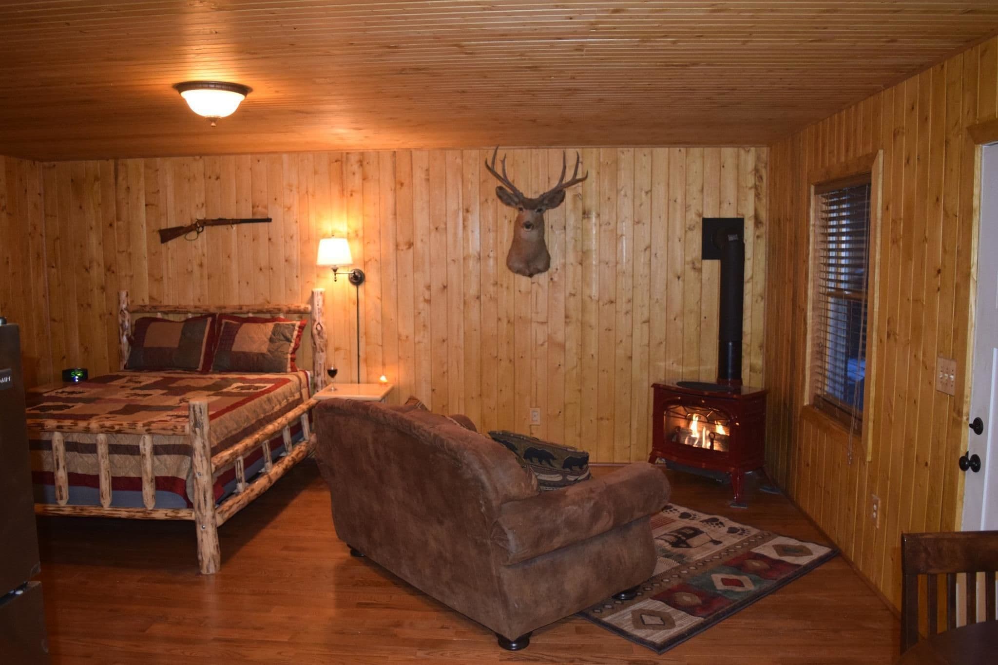 A cozy room with wooden walls, a bed with a patterned blanket, a brown sofa, a mounted deer head, a lamp, and a wood-burning stove.