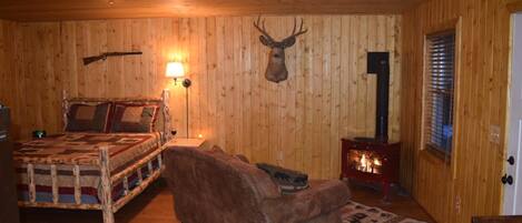 A cozy room with wooden walls, a bed with a patterned blanket, a brown sofa, a mounted deer head, a lamp, and a wood-burning stove.