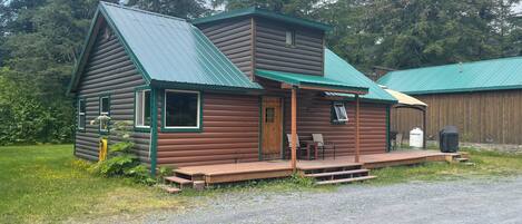 A log cabin with a green roof, a porch with two chairs, and a wooden deck.