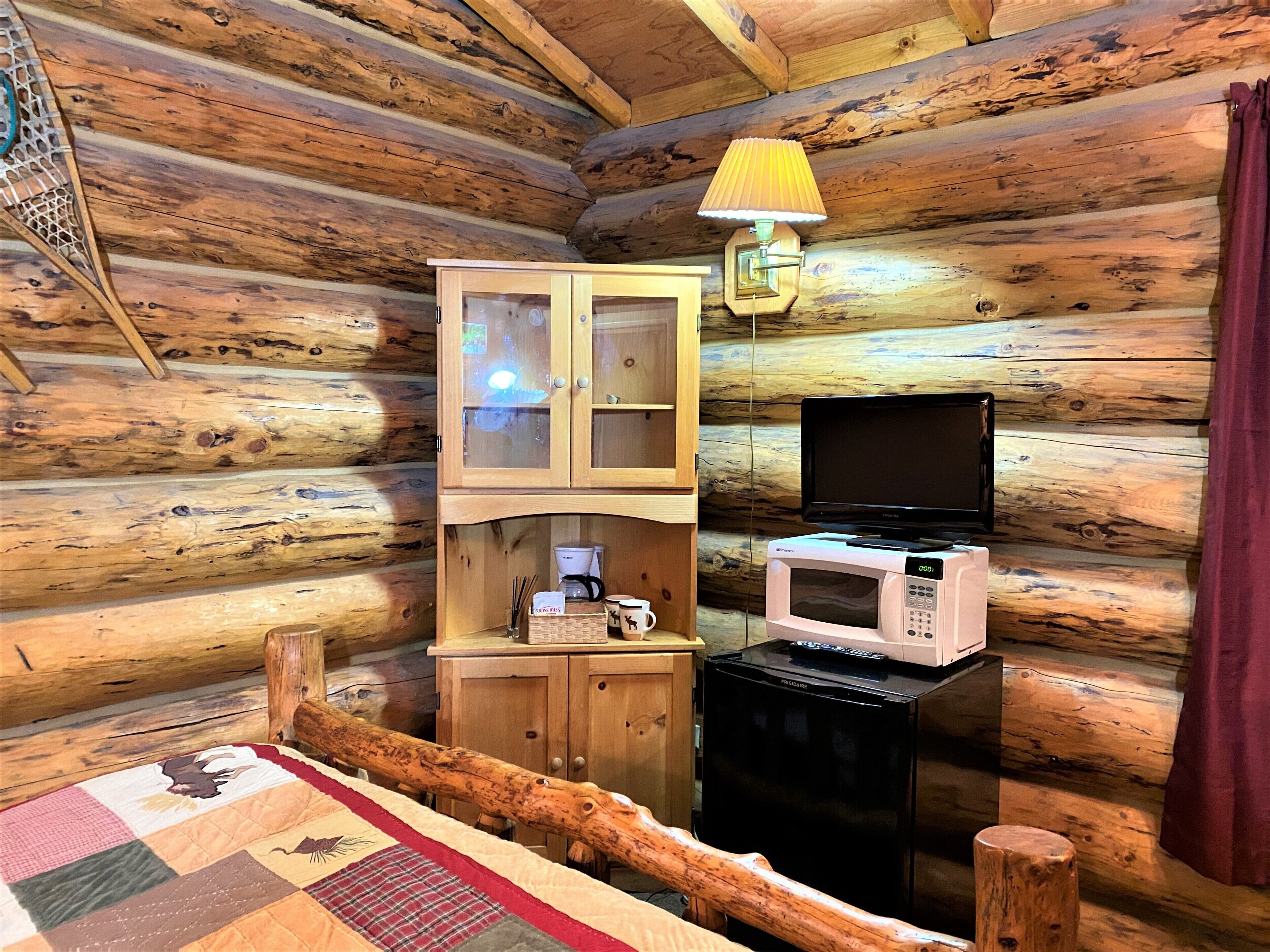 A cozy cabin room with wooden walls, a wooden cabinet with glass doors, a microwave, a television, and a lamp.