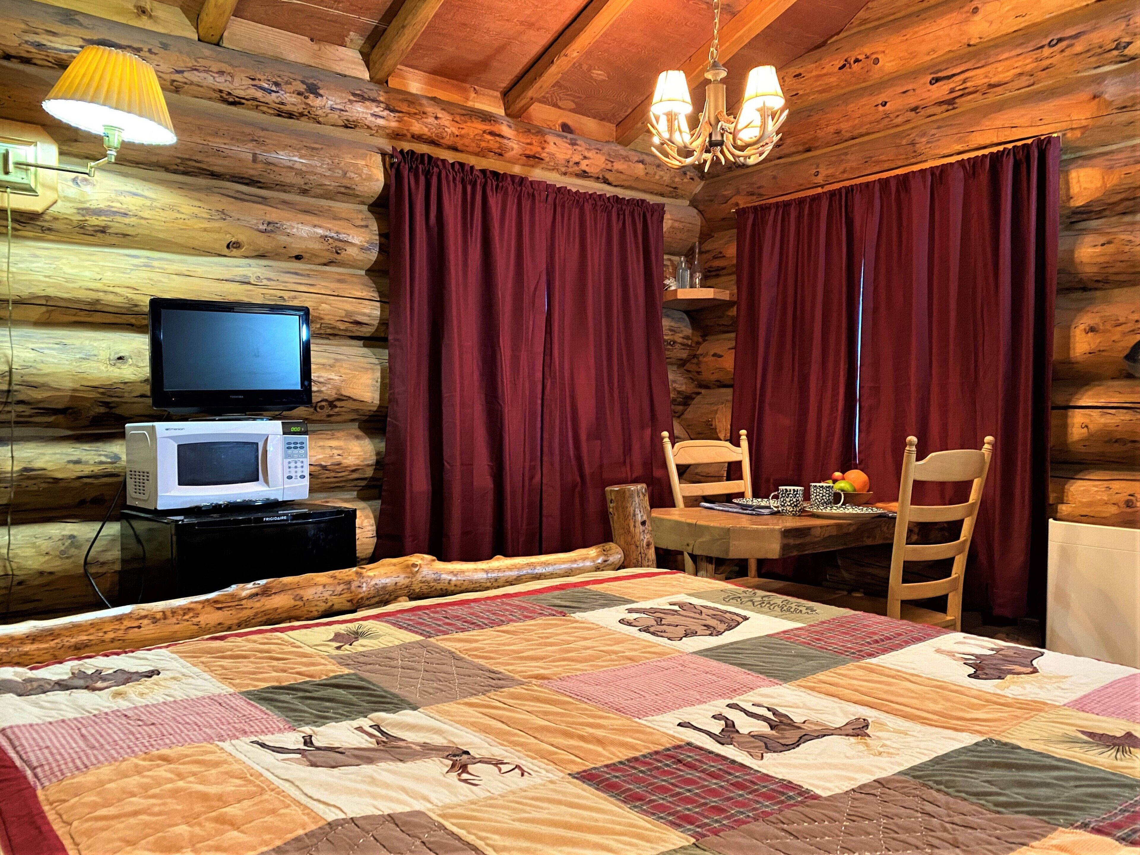 Ein Blockhausszimmer mit einem Bett, einem Tisch mit Stühlen, einem Fernseher, einer Mikrowelle und einer Pendelleuchte.